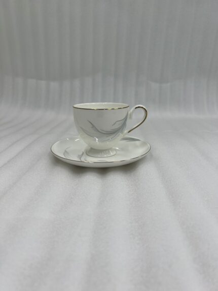 A luxury white super bone china teacup filled with creamy tea, featuring gold-rimmed edges and a delicate grey leaf pattern, resting on a matching saucer on a dark wooden table in a cozy, blurred library background