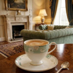A luxury super bone china tea cup with a floral mint-green band and gold rim, filled with coffee, resting on a matching saucer on a polished wooden table in a classic living room with a fireplace
