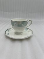 A luxury super bone china tea cup with a floral mint-green band and gold rim, filled with coffee, resting on a matching saucer on a polished wooden table in a classic living room with a fireplace