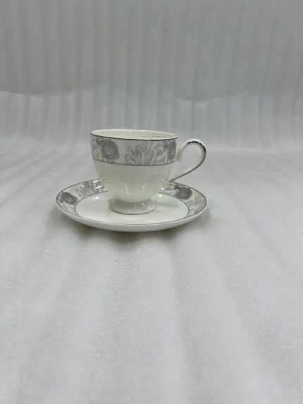 A high-angle shot of a floral-patterned white bone china teacup filled with steaming tea, resting on a matching saucer on a polished wooden table in a luxurious library setting with a green velvet sofa and bookshelves