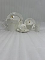 A high-angle shot of a hand pouring hot tea from a white gold-rimmed bone china teapot into a matching cup on a silver tray, surrounded by pastries and a floral centerpiece.