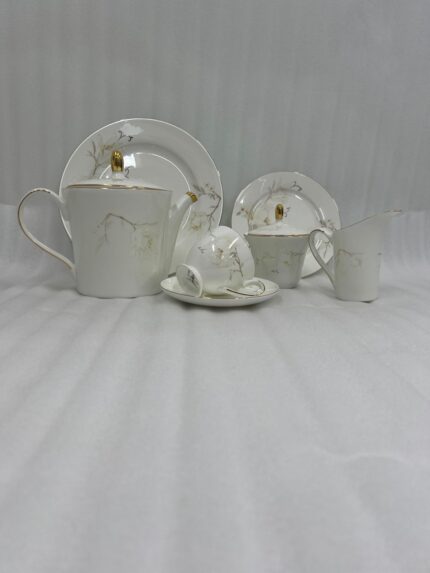 A high-angle shot of a hand pouring hot tea from a white gold-rimmed bone china teapot into a matching cup on a silver tray, surrounded by pastries and a floral centerpiece.