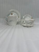 A luxury white Super Bone China tea set with gold rims and a grey feather design, featuring a teapot, creamer, sugar bowl, and cup and saucer on a rustic wooden garden table at sunset