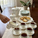 A person pouring milk from a floral bone china creamer into a teacup on a wooden table, surrounded by a matching tea set, teapot, and a plate of sliced cake.