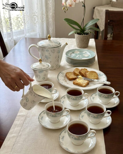 A person pouring milk from a floral bone china creamer into a teacup on a wooden table, surrounded by a matching tea set, teapot, and a plate of sliced cake.
