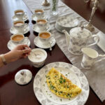 A luxury super bone china teacup and saucer set featuring a grey floral rose pattern, shown on a formal wooden dining table with a matching teapot and breakfast setting.