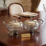 Two-dish Paramount Series stainless steel food warmer with glass bowls and chrome lids, featuring two buffet burners underneath, displayed on a wooden dining table.