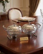 Two-dish Paramount Series stainless steel food warmer with glass bowls and chrome lids, featuring two buffet burners underneath, displayed on a wooden dining table.