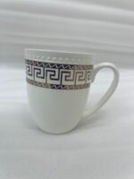 A high-quality white bone china mug filled with steaming milk tea, featuring a gold Greek Key pattern and beaded texture, resting on a rustic wooden garden table