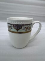 A high-angle close-up of a white bone china mug filled with green tea, featuring an intricate burgundy and gold damask pattern on a mint-green band, resting on a beige linen napkin on a wooden table with books and white rose petals in the background