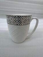 A steaming cup of latte with latte art inside a white bone china mug featuring a gold geometric pattern and quilted texture, resting on a glass saucer with a silver spoon on a wooden table