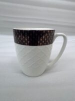 A high-angle shot of a premium white bone china mug with a textured diamond pattern and a dark geometric border, filled with latte art on a wooden table alongside a leather journal and coffee beans