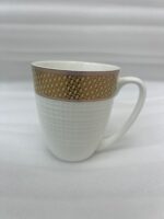 A high-quality bone china coffee mug with a latte inside, featuring a gold woven pattern at the top and an embossed white grid texture on the bottom, set on a dark marble countertop next to coffee beans and a gold spoon