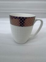 A luxury white bone china mug with a rose gold mesh design on the rim, filled with steaming green tea and a tea leaf floating on top, placed on a textured placemat with a silver spoon on a wooden dining table