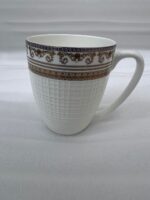 A high-angle shot of a luxury white bone china mug filled with black coffee, featuring an ornate gold Greek key border, placed on a dark wooden table next to a silver spoon in a cozy living room setting