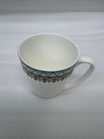 A high-angle close-up of a white bone china mug filled with milk, featuring a gold filigree border and textured lattice design, resting on a rustic blue wooden table in a brightly lit room