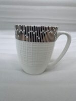 A high-angle shot of a white bone china mug filled with tea, featuring a gold geometric pattern on a wooden outdoor table with a gold spoon and green garden background