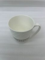 A close-up shot of a white bone china mug with an embossed diamond pattern and a brown Greek key design, filled with latte art, sitting on a granite kitchen countertop next to a silver spoon
