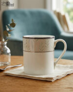 A professional lifestyle shot (3.jpg) showcasing two white Bone China mugs with silver rims and a green botanical leaf pattern, featuring one tall "long" mug and one "short" mug on a linen cloth in a cozy living room setting