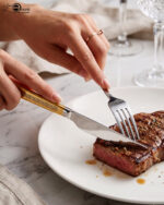 A 36-piece luxury One More cutlery set featuring elegant gold-accented handles with a geometric Greek key pattern, arranged professionally on a dark wood dining table.