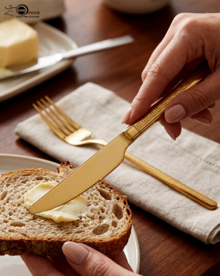 Luxury 36-piece gold-finished cutlery set featuring a geometric Greek key handle design, displayed elegantly on a dark grey placemat.