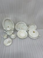 An elegant white super bone china dinner set featuring delicate beige floral patterns and gold rims, arranged on a textured beige surface. The set includes plates, bowls, a teapot, a covered casserole dish, a cream pitcher, a sugar bowl, and a teacup with a saucer.
