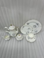 A luxury white super bone china tea set with grey floral patterns and gold rims on a wooden table, featuring a teapot, teacup, plates, and a steaming cup of tea against a snowy window BG.