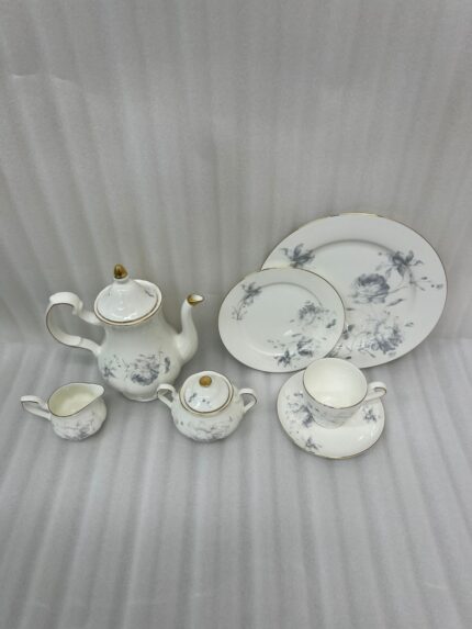 A luxury white super bone china tea set with grey floral patterns and gold rims on a wooden table, featuring a teapot, teacup, plates, and a steaming cup of tea against a snowy window BG.