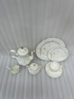 A luxury white super bone china tea set with grey floral patterns and gold rims on a wooden table, featuring a teapot, teacup, plates, and a steaming cup of tea against a snowy window BG.