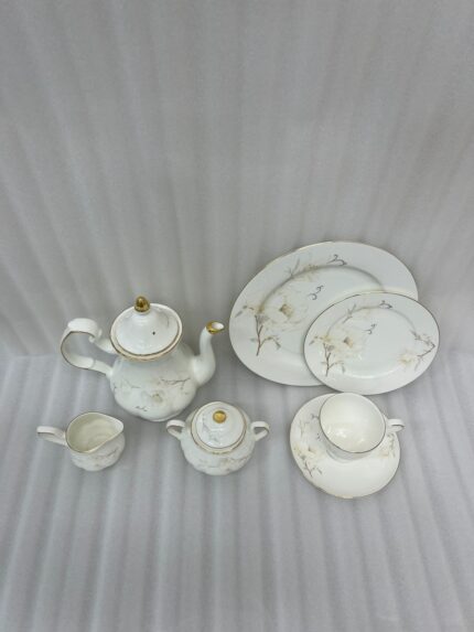 A luxury white super bone china tea set with grey floral patterns and gold rims on a wooden table, featuring a teapot, teacup, plates, and a steaming cup of tea against a snowy window BG.