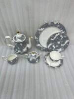 A premium grey and white floral pattern super bone china tea set featuring a teapot with gold accents, a creamer, a sugar bowl, plates, and a filled teacup with a saucer on a wooden table.
