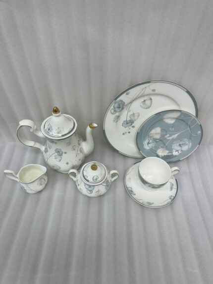 A premium super bone china tea set featuring a classic teapot, sugar bowl, creamer, teacup with saucer, and matching plates, all adorned with an elegant light blue and grey floral pattern on a wooden table.