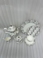A premium bone china tea and coffee set with a grey floral pattern and silver rims on a dark wooden table. The collection includes a teapot, creamer pitcher, sugar bowl, plates, and a steaming cup of black coffee on a saucer with a silver spoon, set against a bright window showing a lush garden background