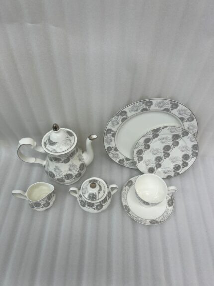 A premium bone china tea and coffee set with a grey floral pattern and silver rims on a dark wooden table. The collection includes a teapot, creamer pitcher, sugar bowl, plates, and a steaming cup of black coffee on a saucer with a silver spoon, set against a bright window showing a lush garden background
