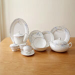 A comprehensive 61-piece super bone china dinner set for an 8-person serving, displayed on a wooden table against a neutral background. The white ceramic tableware features a delicate light blue and grey floral pattern with thin silver rims. The set includes a large oval platter, dinner plates, side plates, soup bowls, a covered casserole dish, a teapot, a creamer, a sugar bowl, and tea cups with saucers.