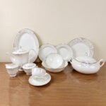 Detailed product photograph of an elegant 61-piece super bone china dinner set for 8-person serving, displayed on a polished wooden surface. The set includes a full tea service (teapot, creamer, sugar bowl, teacup and saucer), multiple plates and bowls, a large lidded tureen, and various serving pieces, all featuring delicate light gold damask filigree patterns and gold trim on a cream-white surface. The 'one more' brand logo is visible in the upper left corner.