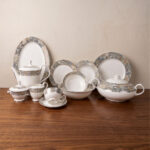 A photograph of a One More Super Bone China 61-piece dinner set arranged on a dark walnut wood table against a neutral beige wall. The luxurious set, designed for 8-person serving, features white bodies and elegant grey-gold marble-pattern rims with subtle gold detailing. It displays a variety of plates, a matching teapot, a large covered tureen, open serving bowls, a cream pitcher, and teacups with saucers. The brand logo is visible in the top left corner
