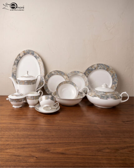 A photograph of a One More Super Bone China 61-piece dinner set arranged on a dark walnut wood table against a neutral beige wall. The luxurious set, designed for 8-person serving, features white bodies and elegant grey-gold marble-pattern rims with subtle gold detailing. It displays a variety of plates, a matching teapot, a large covered tureen, open serving bowls, a cream pitcher, and teacups with saucers. The brand logo is visible in the top left corner