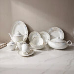 A detailed product photograph of a 61-piece 'super bone' china dinner set by One More, arranged on a white marble countertop against a neutral background. The comprehensive set, with service for 8 people, includes a teapot, creamer, sugar bowl with lid, tea cup with saucer, a large lidded casserole dish, numerous plates (dinner, salad, soup) and bowls stacked, and an oval serving platter. Each piece features a matching, delicate blue and grey floral motif with a refined gold rim, presented under elegant natural lighting. The One More logo is in the upper left