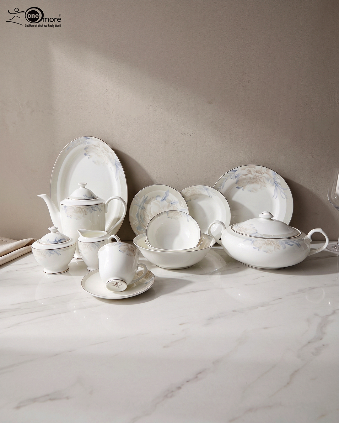 2 A detailed product photograph of a 61-piece 'super bone' china dinner set by One More, arranged on a white marble countertop against a neutral background. The comprehensive set, with service for 8 people, includes a teapot, creamer, sugar bowl with lid, tea cup with saucer, a large lidded casserole dish, numerous plates (dinner, salad, soup) and bowls stacked, and an oval serving platter. Each piece features a matching, delicate blue and grey floral motif with a refined gold rim, presented under elegant natural lighting. The One More logo is in the upper left