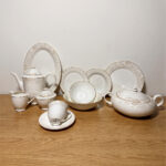 A comprehensive collection of 61 pieces of white bone china dinnerware featuring an intricate gold and cream floral pattern with gold rims, arranged elegantly on a wooden table.