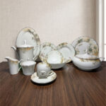 A premium 61-piece white super bone china dinner set with a delicate green and gold floral pattern displayed on a polished dark wood table. The set features an elegant teapot, casserole tureen, serving bowls, large oval platter, dinner plates, side plates, and tea cups with saucers against a neutral textured background