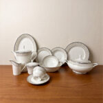 An elegant 61-piece white super bone china dinner set arranged on a polished wooden table against a neutral beige wall. The porcelain tableware features sophisticated silver-grey textured borders and includes dinner plates, bowls, a large oval serving platter, a covered soup tureen, and a complete tea service with a teapot, creamer, sugar bowl, cups, and saucers