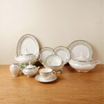 An elegant 61-piece super bone china dinnerware set for an 8-person serving, featuring white plates, bowls, a teapot, a covered tureen, and teacups with detailed beige and silver patterned rims, neatly arranged on a wooden table