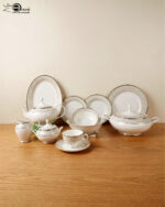 An elegant 61-piece super bone china dinnerware set for an 8-person serving, featuring white plates, bowls, a teapot, a covered tureen, and teacups with detailed beige and silver patterned rims, neatly arranged on a wooden table