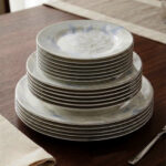 A close-up photograph of a massive, multi-tiered stack of sophisticated grey and white dinner plates featuring a detailed peony floral design with blue accents and silver rims. The plates are neatly arranged on a grey linen runner on a dining table, next to a crystal wine glass and another set of plates. The brand "One More" logo is visible in the background
