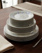 A close-up photograph of a massive, multi-tiered stack of sophisticated grey and white dinner plates featuring a detailed peony floral design with blue accents and silver rims. The plates are neatly arranged on a grey linen runner on a dining table, next to a crystal wine glass and another set of plates. The brand "One More" logo is visible in the background