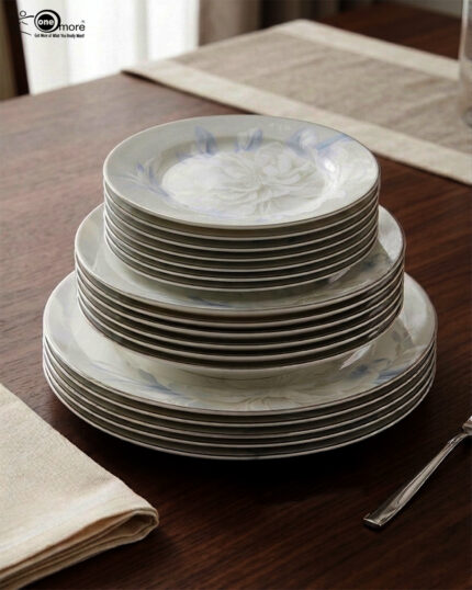 A close-up photograph of a massive, multi-tiered stack of sophisticated grey and white dinner plates featuring a detailed peony floral design with blue accents and silver rims. The plates are neatly arranged on a grey linen runner on a dining table, next to a crystal wine glass and another set of plates. The brand "One More" logo is visible in the background