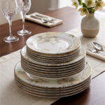 A beautifully stacked 18-piece premium Super Bone China plate set featuring an elegant floral pattern and fine trim, arranged on a beige linen table runner alongside crystal wine glasses and a rustic flower vase.