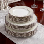 A professionally styled stack of the OneMore 18-piece super bone china dinnerware set, showcasing six quarter plates on top, six deep plates in the middle, and six large rice plates at the bottom, featuring an elegant light-green patterned border on a silver table runner